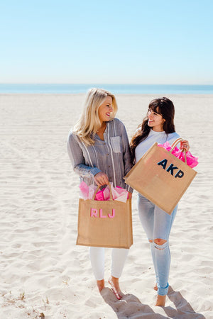 Two women on the beach hold monogrammed jute bags with pink tissue paper inside.