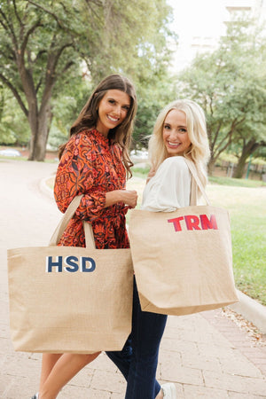 A blonde and a brunette smile while holding monogrammed jute tote bags