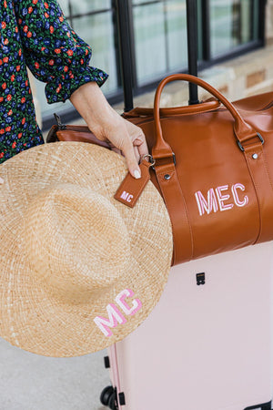 A person attaches a straw beach hat to a tan duffel bag with a tan leather hat clip which has a pink monogram.