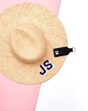 A black leather hat clip with a beige monogram attaches to a straw hat.