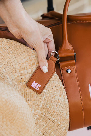 A person attaches a straw beach hat to their duffel with a tan leather hat clip with a pink monogram.