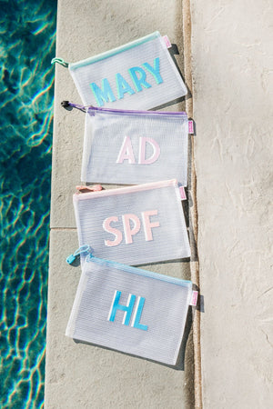 A group of white pool bags with pastel colored zippers and monograms are laid out next to a pool.