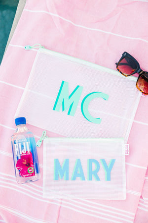A small and a large pool bag are laid on a pink towel with a water bottle and some sunglasses