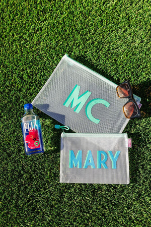 A small and a large pool bag are laid on the grass with a water bottle and some sunglasses