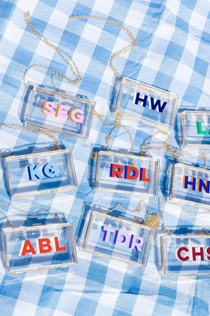 Clear stadium bags with gold chain straps, each with bold shadow monograms over blue-white gingham for a custom gameday look