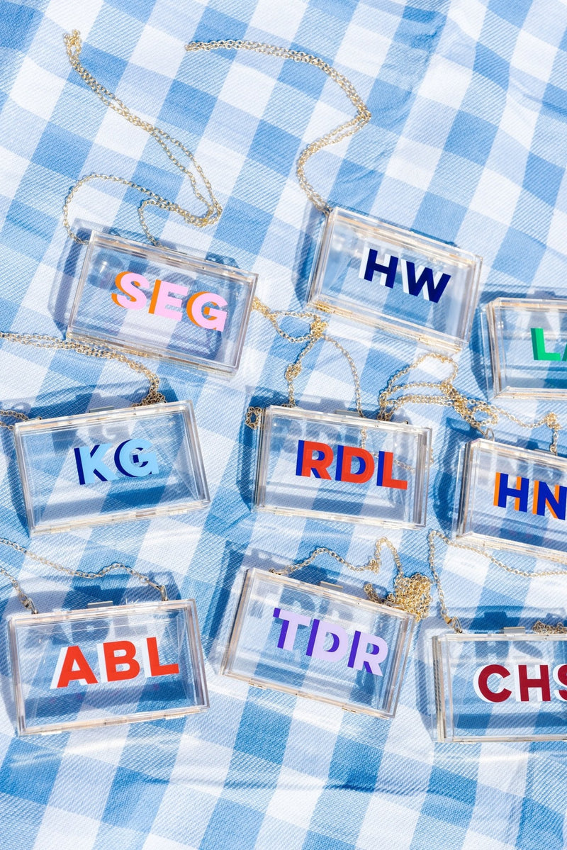 Clear stadium bags with gold chain straps, each with bold shadow monograms over blue-white gingham for a custom gameday look