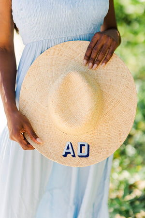 A woman in a blue dress holds a straw hat with "AD" embroidered on the brim in blue