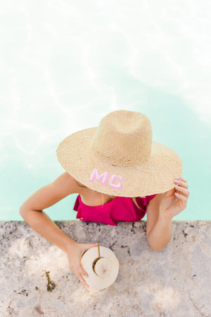 A woman in a pink swimsuit wears a monogrammed straw beach hat