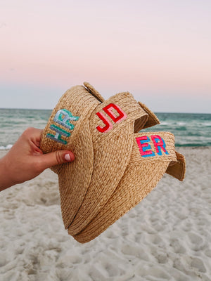 A person holds up a group of straw visor with different colored embroidered monograms