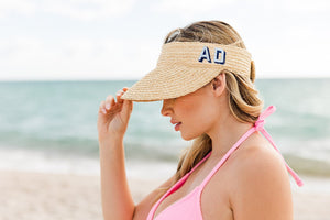 A girl on the beach holds onto her straw visor with a blue monogram.