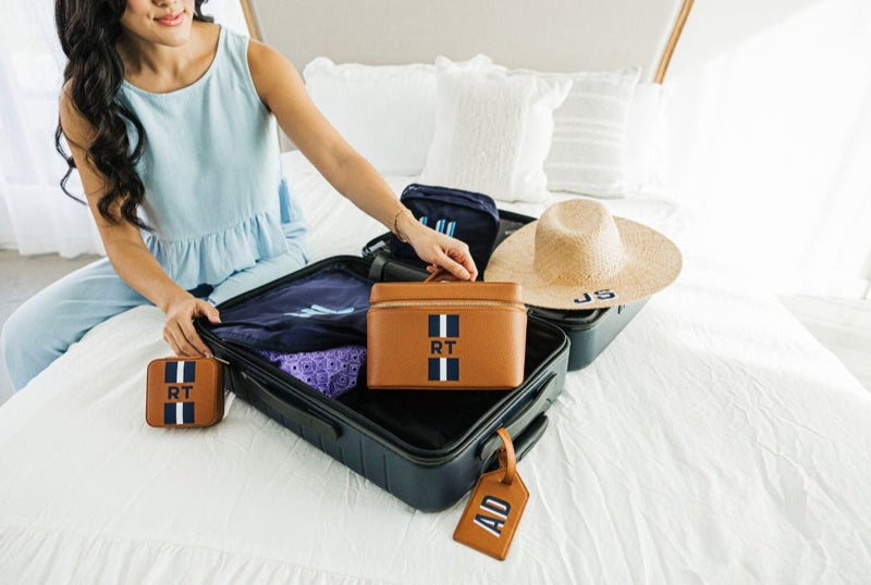 A person packs a suitcase with personalized products like a leather train case and jewelry case with navy stripe monograms.