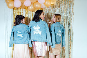 Three girls celebrate by wearing customized jean jackets that say "The Party" in white.