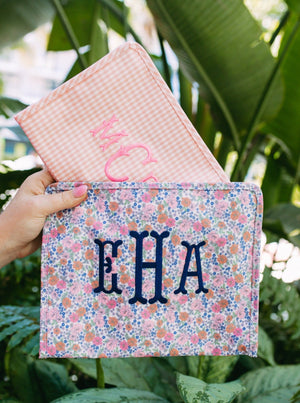 A person holds up a pink and a floral patterned pouch with monograms in front of some greenery.