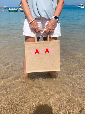 A person in the water holds up a cotton tote which reads "VACAY" across the front in pink and red letters