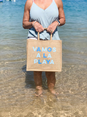 A person in the water holds up a cotton tote which reads "VAMOS A LA PLAYA" across the front in blue letters