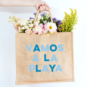 A jute bag is customized with "Vamos A La Playa" in alternating blue font.
