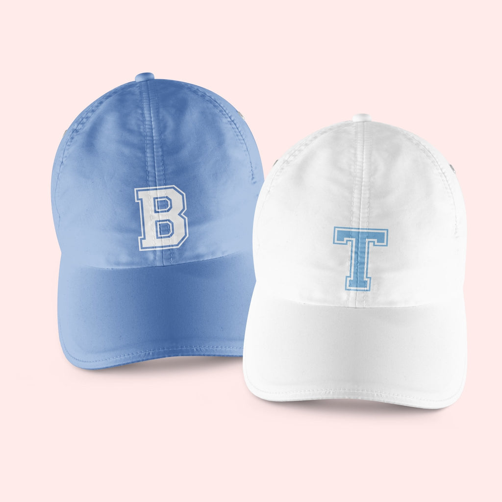 A blue and white baseball hat are customized with varsity letter style monograms.