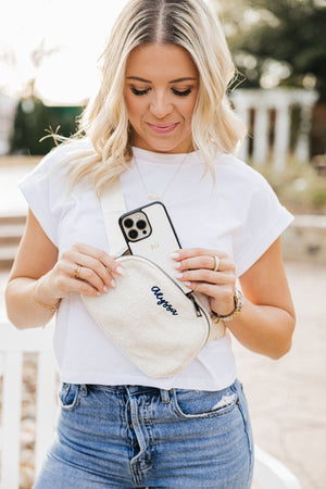 A woman in a white t shirt puts her phone customized with a gold monogram into her belt bag