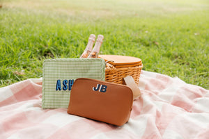 A monogrammed, tan vegan leather pouch sits on a picnic blanket