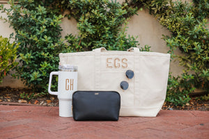 A black vegan leather pouch with a gold foil monogram sits with a monogrammed tumbler and canvas tote