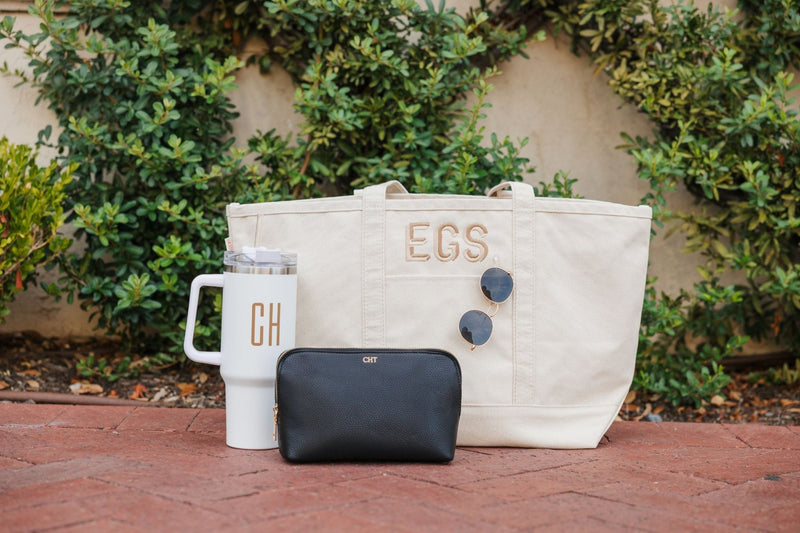 A black vegan leather pouch with a gold foil monogram sits with a monogrammed tumbler and canvas tote