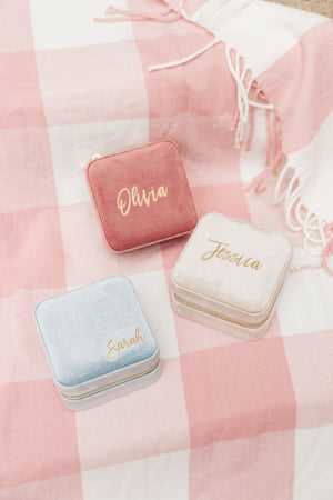 Three velvet jewelry cases in pink, blue, and white sit on a pink plaid blanket