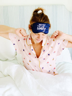 A girl stretches in her bed wearing a navy "Wake Me In Italy" sleep mask on her head.