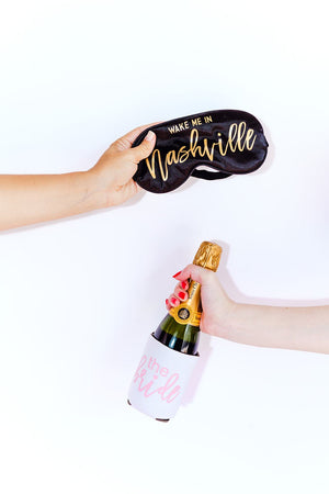 A person holds up a customized black sleep mask while another person holds up a bottle of champagne with a personalized can cooler.