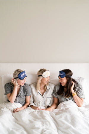 Three girls sit in a bed wearing customized sleep masks.