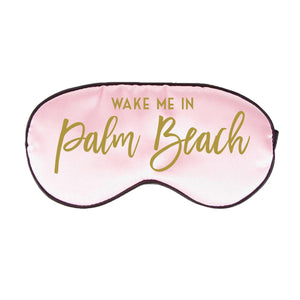 A pink sleep mask is customized with a gold script font which reads "Wake Me In Palm Beach".
