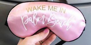 A pink sleep mask is customized with a gold and white font which reads "Wake Me In Palm Beach".