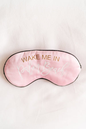 A pink sleep mask is customized with a gold and white script font which reads "Wake Me In Palm Beach".
