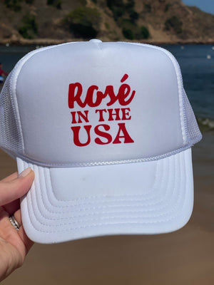 A woman at the beach holds up a white trucker hat that says "Rosé In The USA"